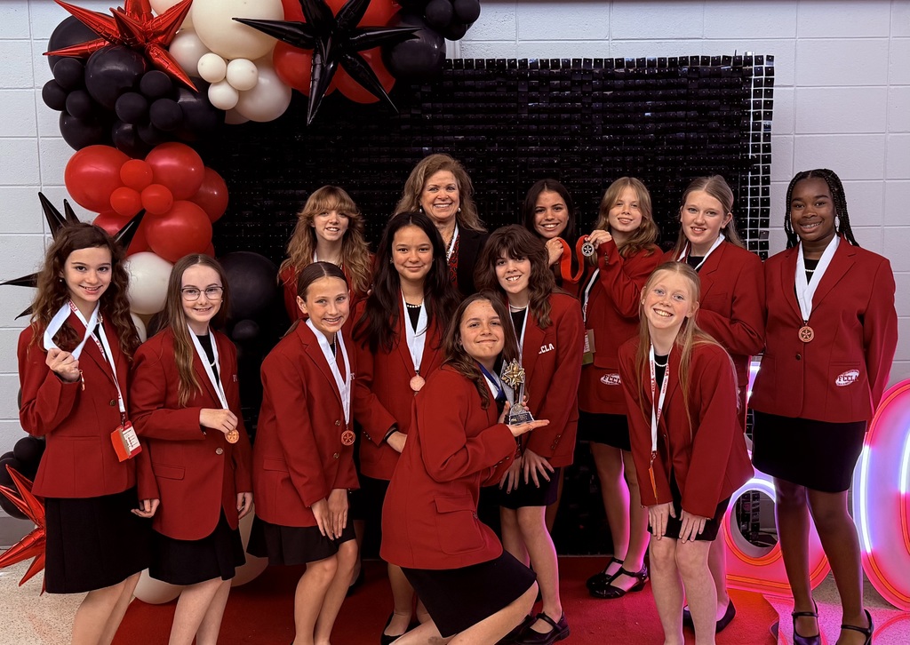 12 female students and 1 teacher is red blazers and black skirts posing in front of red, black, and white balloons