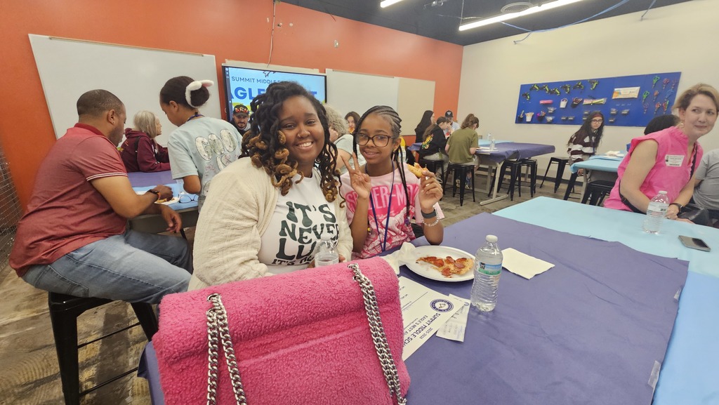 Student holds up a peace sign and smiles while sitting next to her mom and enjoying a slice of pizza.