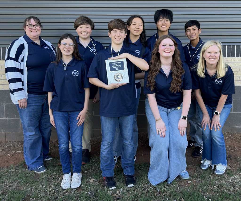 Seven 7th/8th graders in navy shirts and jeans posing with two female teachers. The student in the middle of the front row is holding a plaque