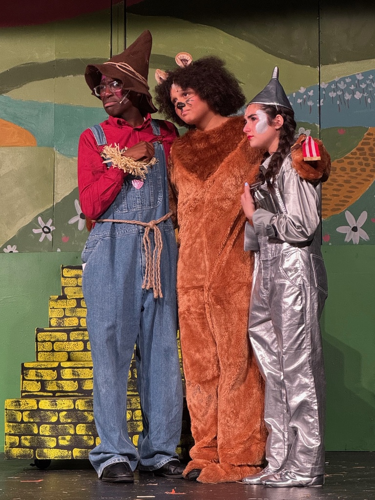 The Scarecrow, the Cowardly Lion, and the Tin Man on stage with a yellow brick road behind them