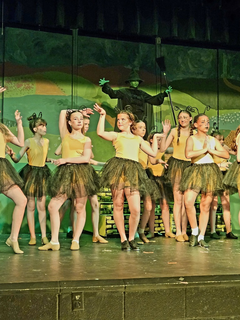 A group of girls in yellow tanks and dark green tutus with a Wicked Witch behind them