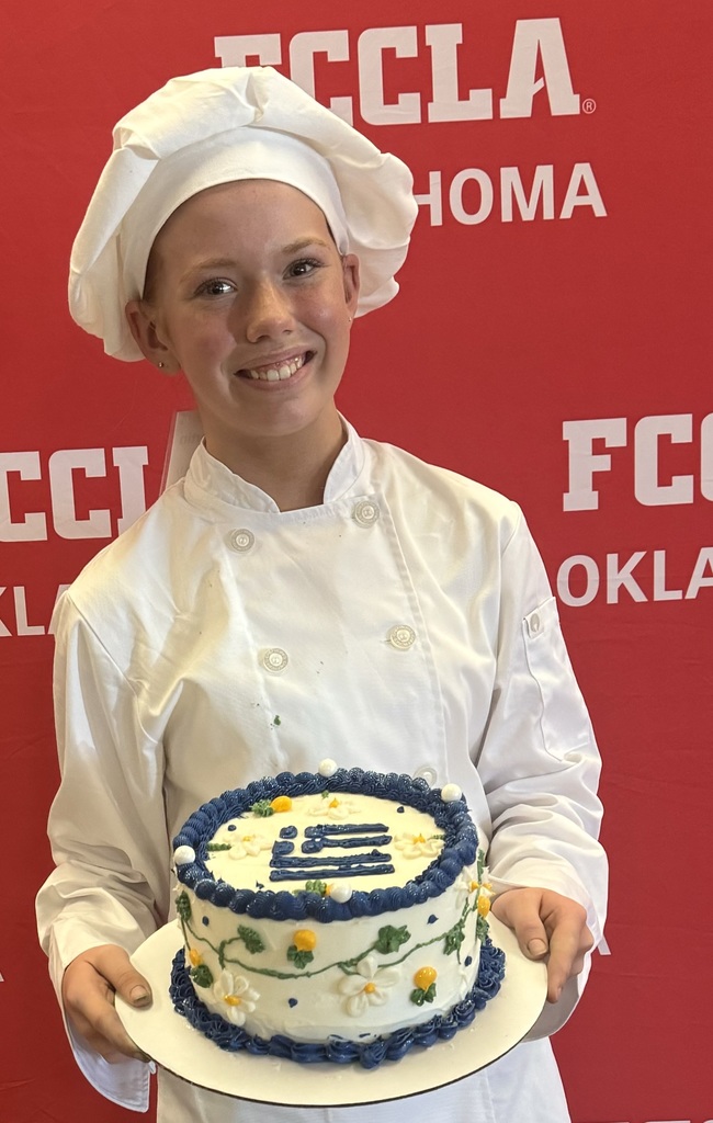 Elizabeth is standing in front of a red FCCLA sign while holding the cake she decorated to win 1st place at the state competition. 