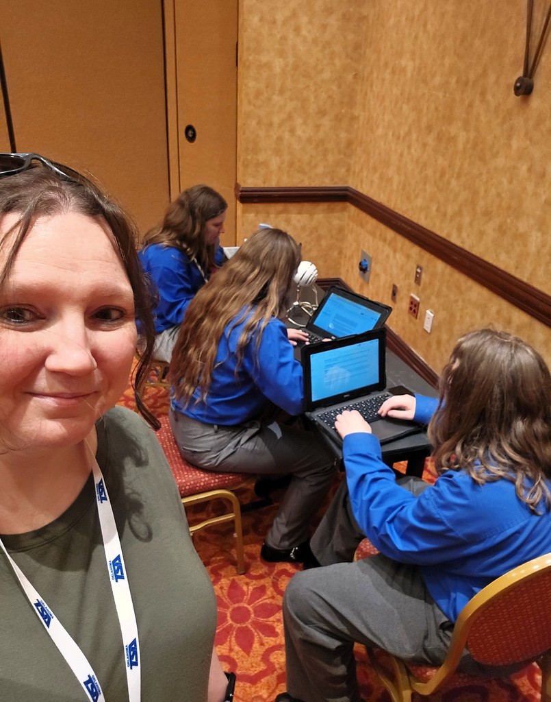 TSA Advisor, Stacie Andrews, smiles for the camera while TSA student members can be seen sitting and working on laptops in the background.