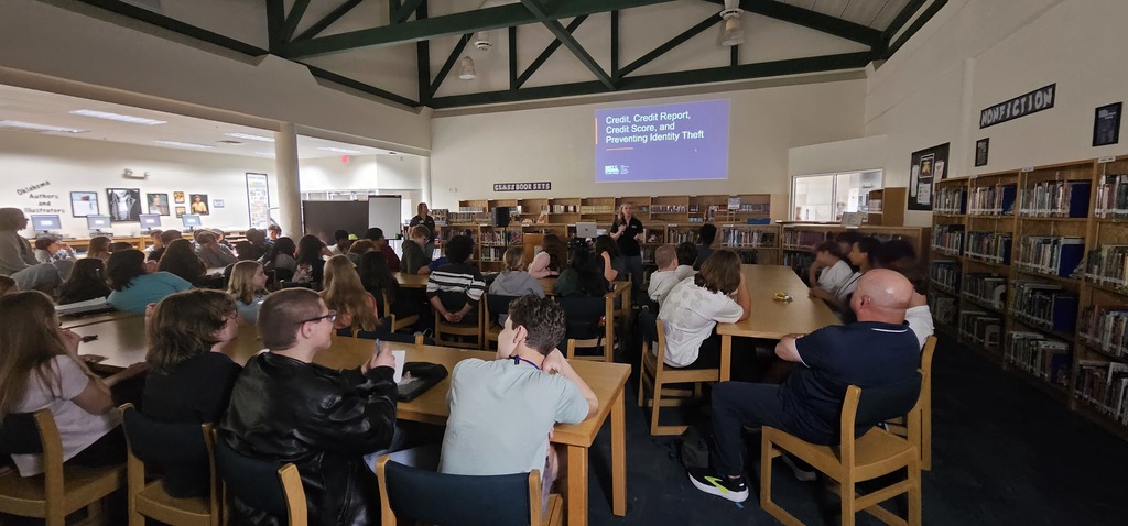 Students sitting in tables watching two ladies present with a slideshow projected onto the wall behind the presenters