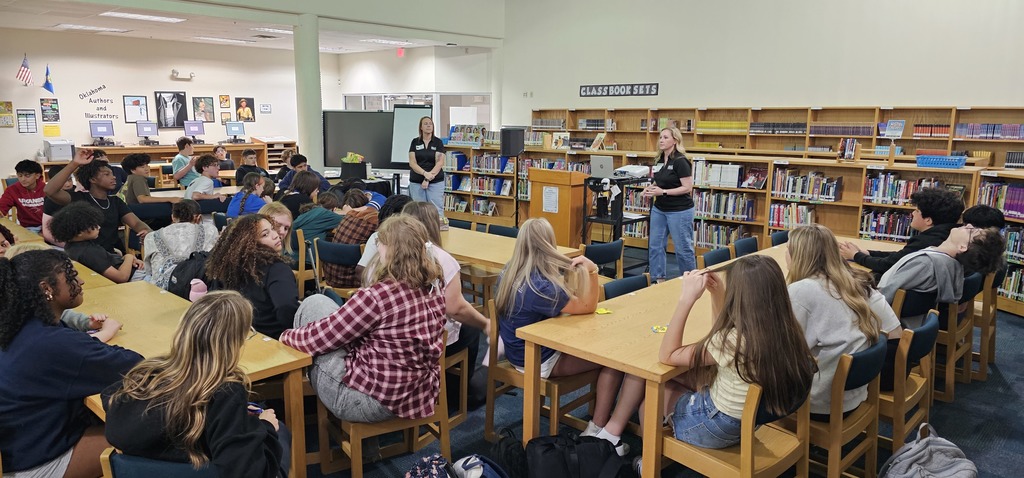 Students sitting in tables in the Media Center listening to two adult women in jeans and black shirts.