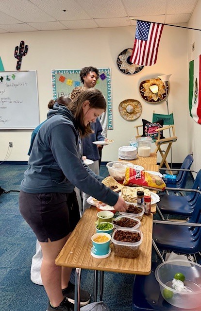 A table with meat, cheese, sour cream, tacos, chips, and plates. Students are standing in front of the table adding food to their plates