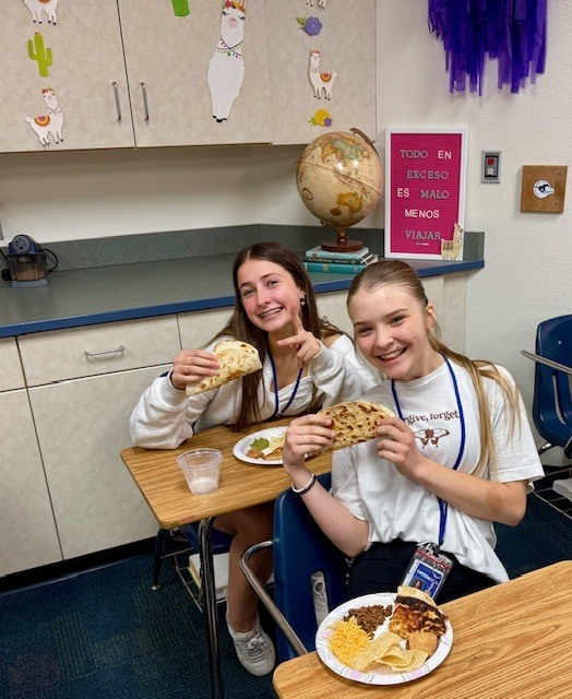 Two teens sitting at desks eating Mexican food and posing holding tacos