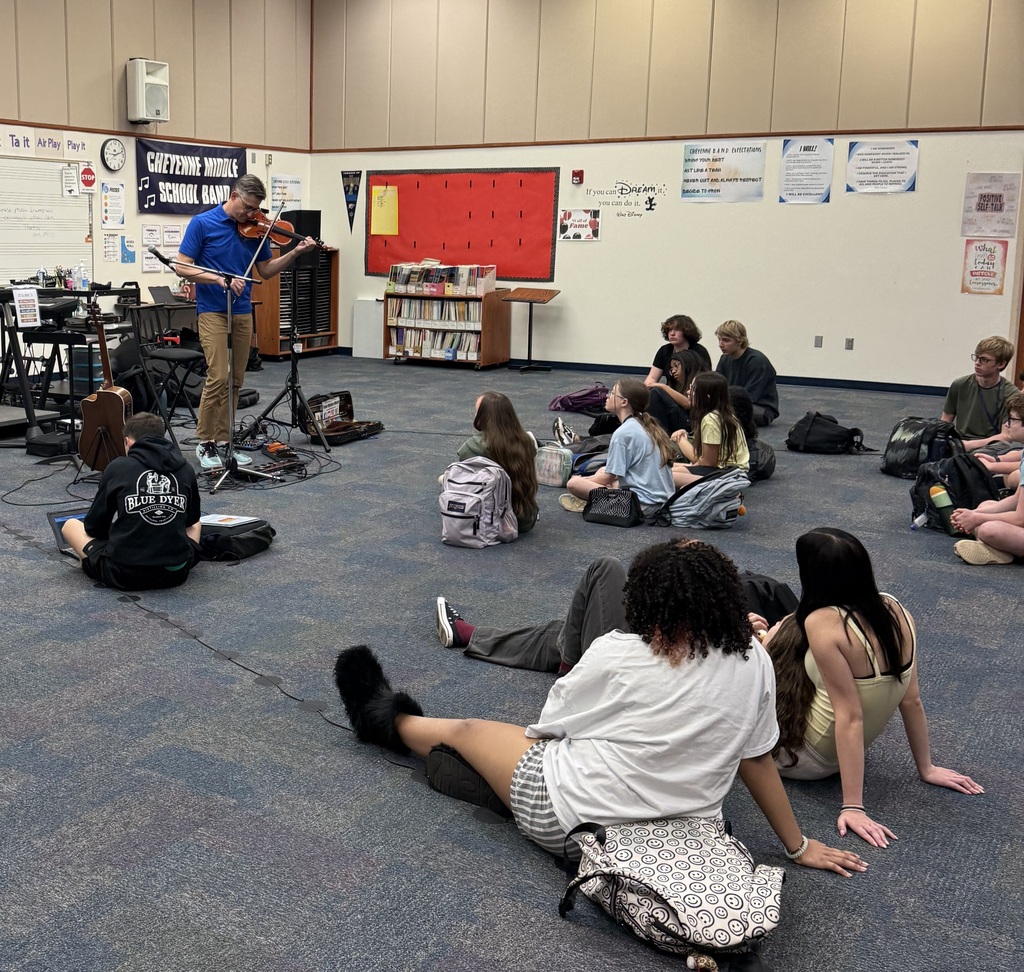 A man in a blue shirt playing the violin while teens sit on the floor watching