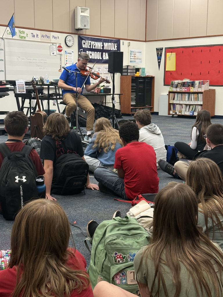 A man in a blue shirt playing the violin while students sit on the floor watching him perform