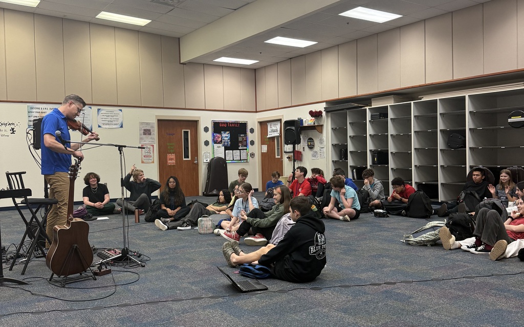 A man in a blue shirt playing the violin while students sit on the floor watching him perform. There is a guitar on a stand next to the man.