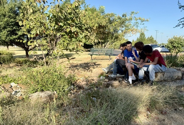 5 teen boys sitting on some rocks outdoors talking with one another.