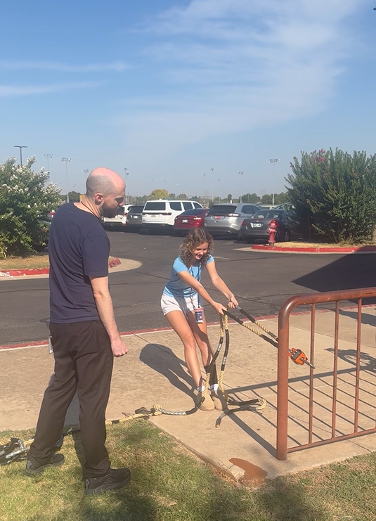 A teen girl in a blue shirt pulling on a crane scale while a male teacher reads the measurement
