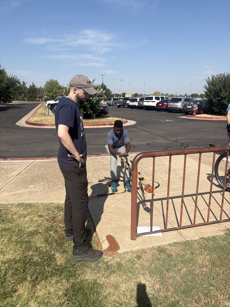 A teen boy pulls on a crane scale while a male teacher reads the measurement