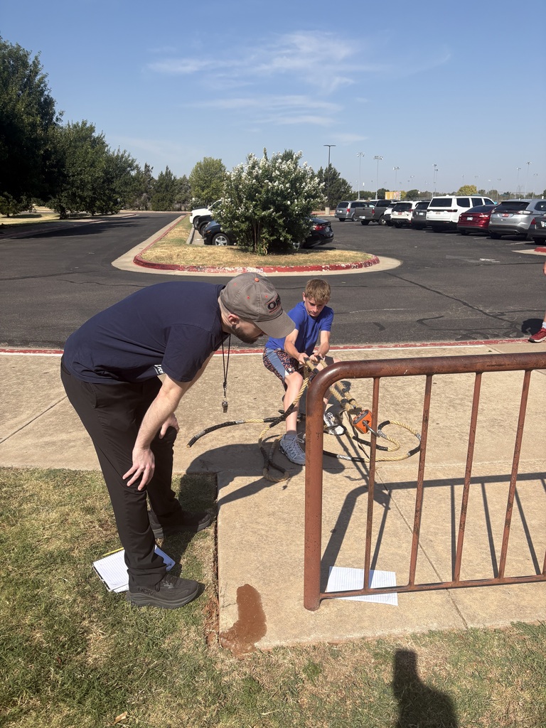 A teen boy pulls on a crane scale while a male teacher reads the measurement