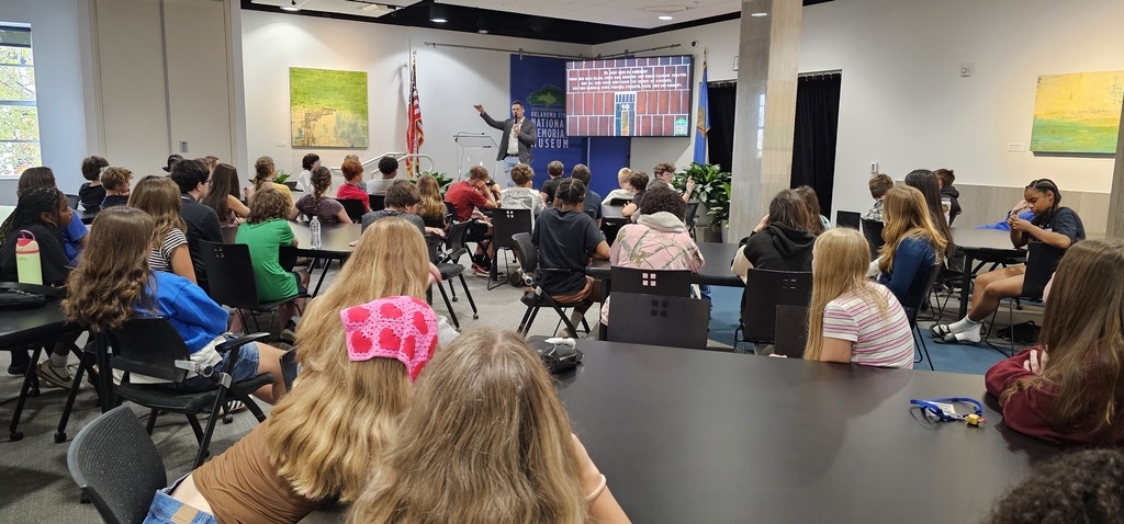A group of students sitting at black tables listening to a male speaker present with a TV screen behind him and an American flag to his right.