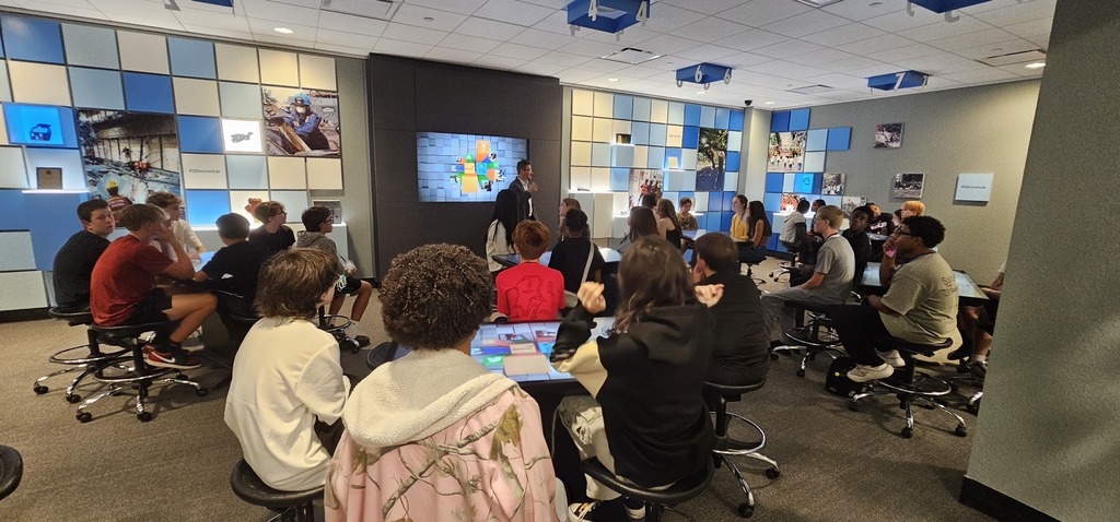 Students sitting around touch screen computer panels with a male speaker presenting in a room with light blue, royal blue, and white squares behind him