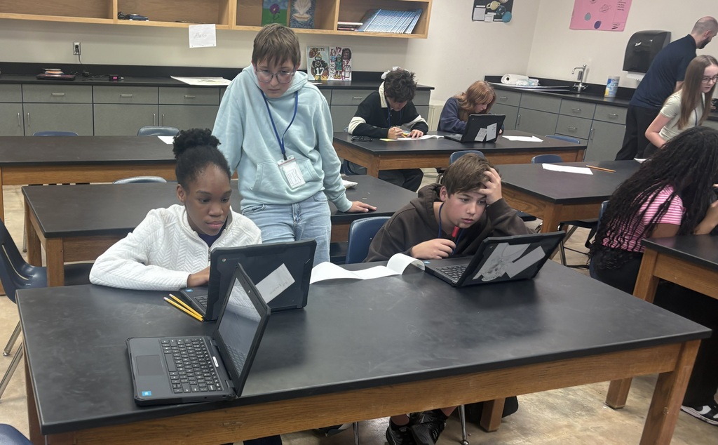 A girl and boy researching on their chromebooks, while a boy in a blue hoodie stands behind them looking on. There is a sheet of white paper on the table between the two students.