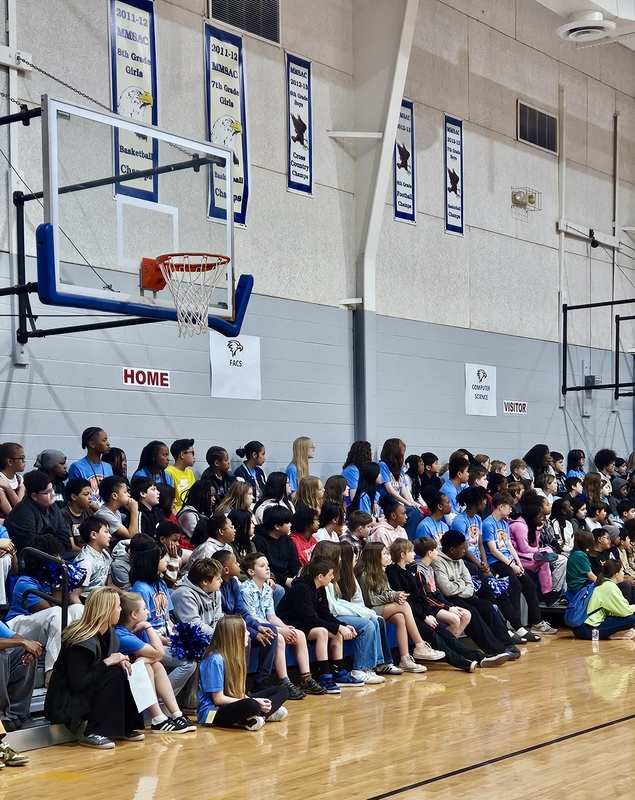Large group of 5th grade students, WEB leaders, and various teachers sit in the bleachers listening to a speaker during 5th grade visits.