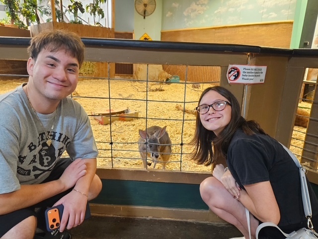 two students posing by a wire gate with a baby kangaroo behind it