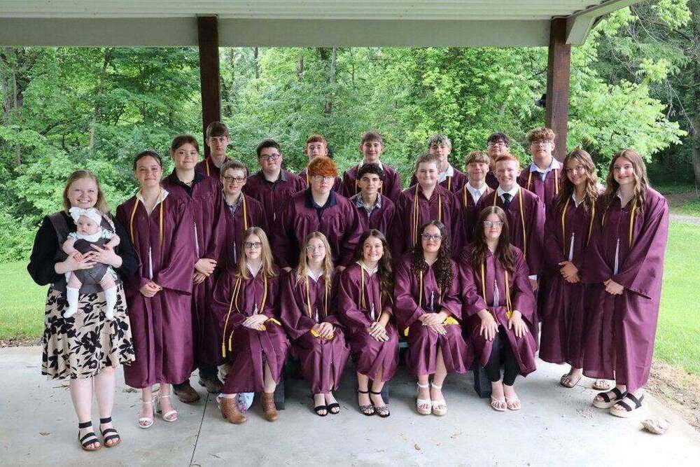 A group photo of graduates in maroon gowns and yellow stoles posing under an outdoor pavilion. A woman holding a baby stands to the left.