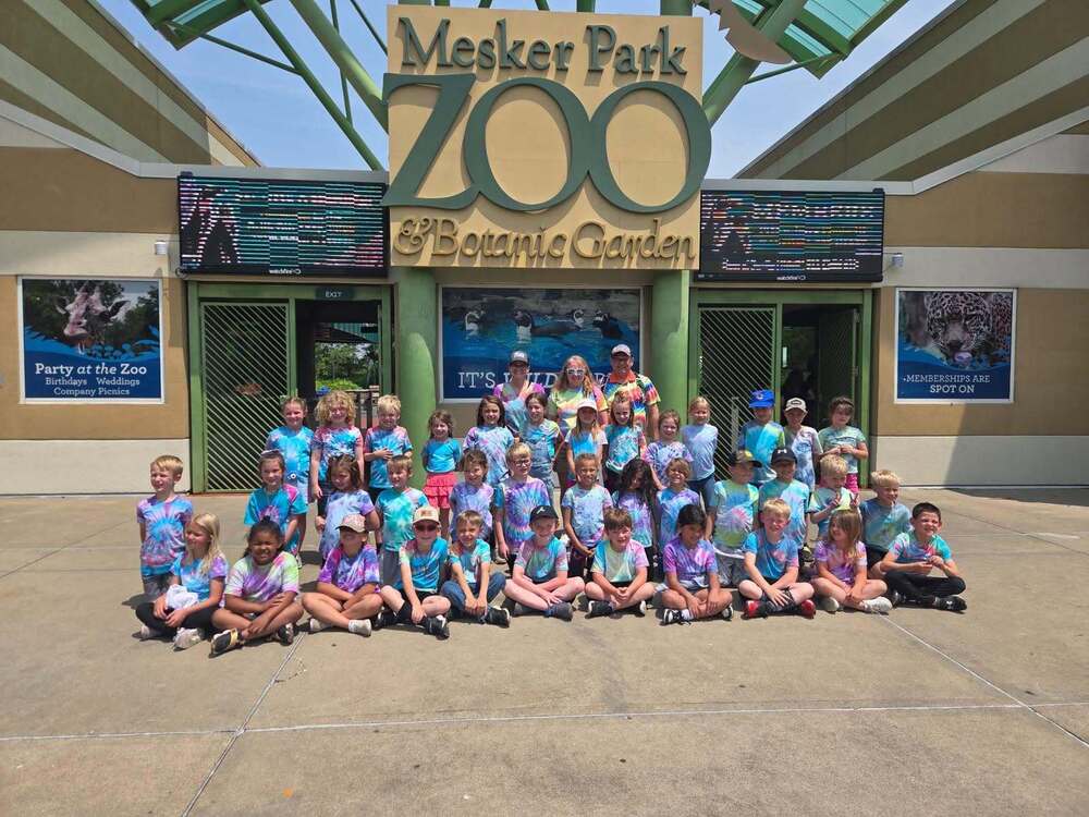 A large group of children and a few adults posing for a photo in front of the entrance to the Mesker Park Zoo & Botanic Garden. Everyone in the group is wearing matching bright tie-dye shirts.