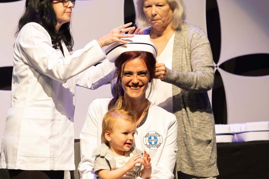 smiling nursing student holding her baby while two staff pin on the nurse cap