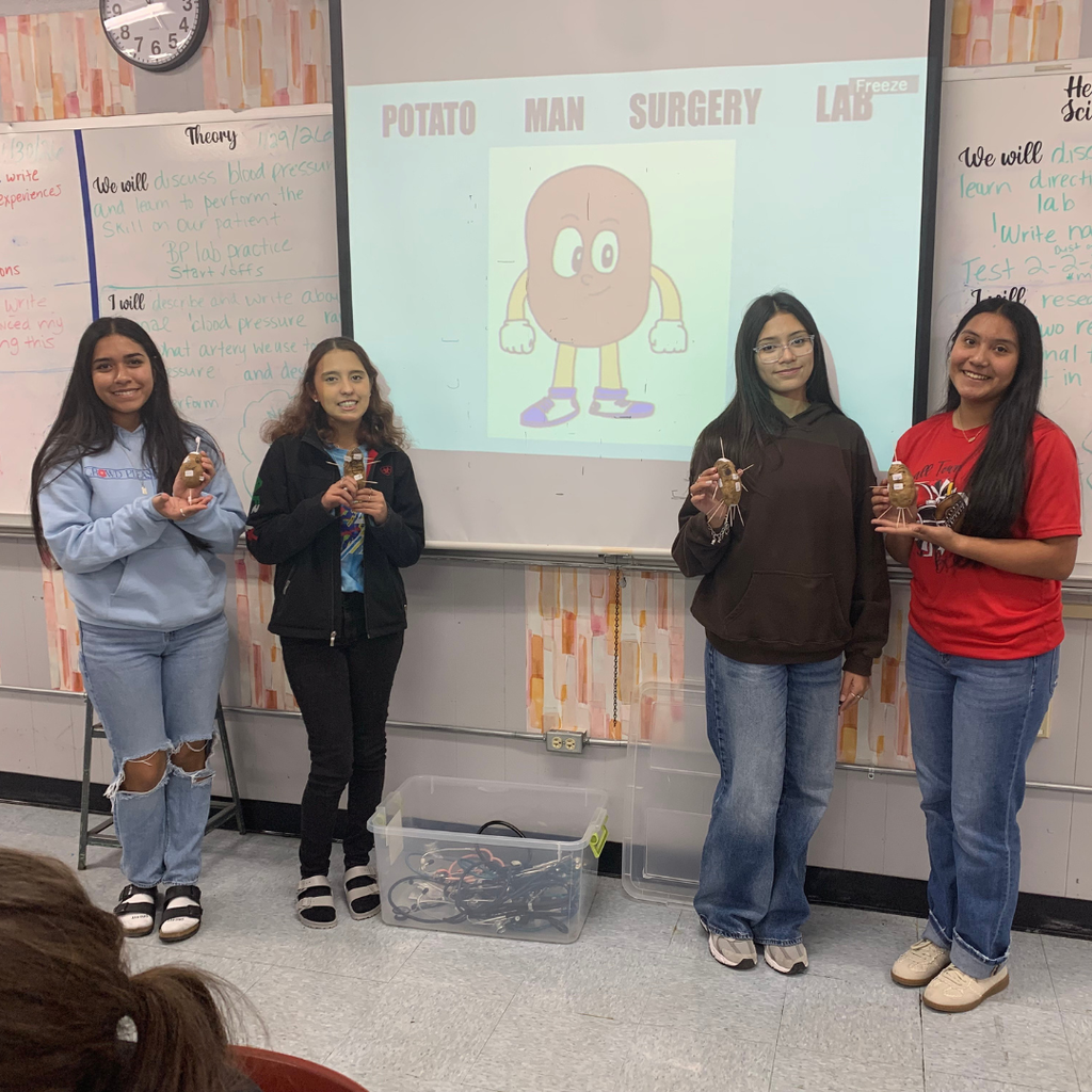 Four female students standing in a classroom holding up their 'Potato Man' projects in front of a whiteboard labeled 'Potato Man Surgery Lab'