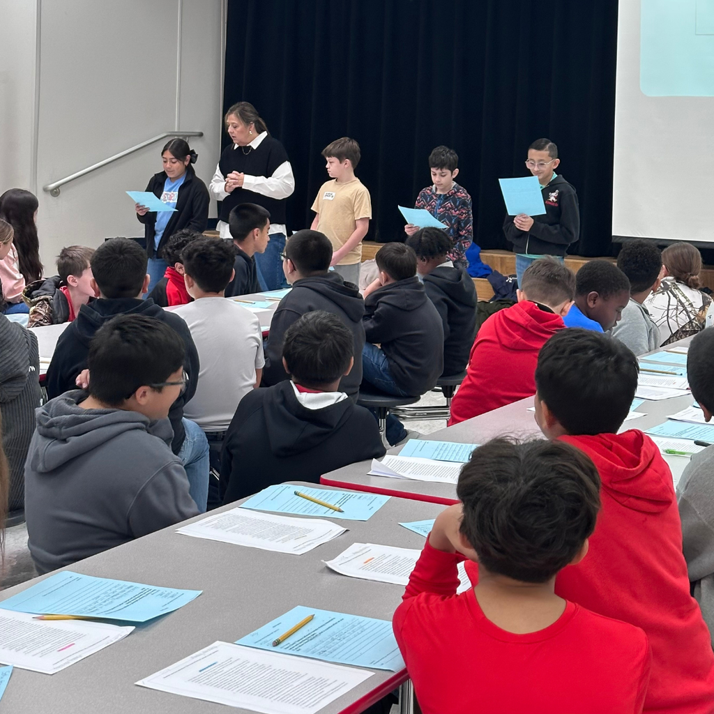 A group of 5th-grade students at Northside Elementary sit at long tables in the cafeteria, focused on a writing workshop. A teacher stands among them, guiding the lesson as students look at their papers and interact.