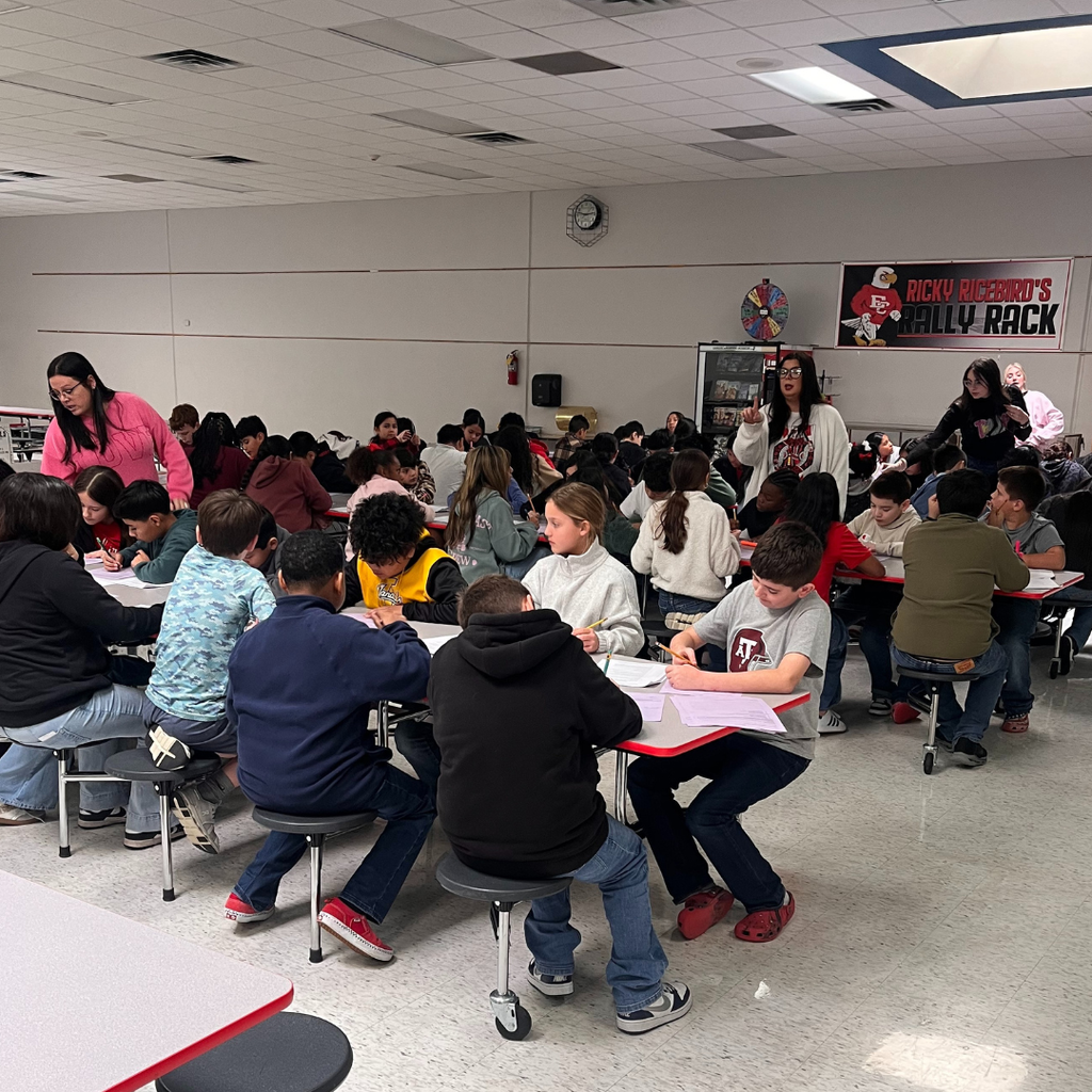 A group of 5th-grade students at Northside Elementary sit at long tables in the cafeteria, focused on a writing workshop. A teacher stands among them, guiding the lesson as students look at their papers and interact.