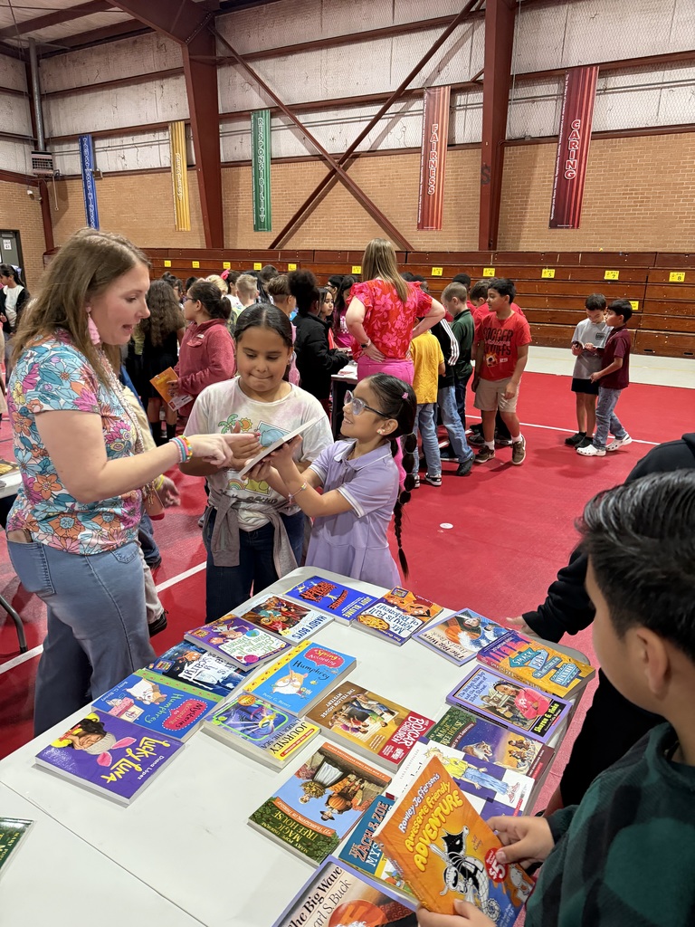 A teacher and a group of elementary students gather around a table covered in a variety of colorful children's books. One student is smiling as she looks at a book she just picked out during the Chase’s Chapters event.