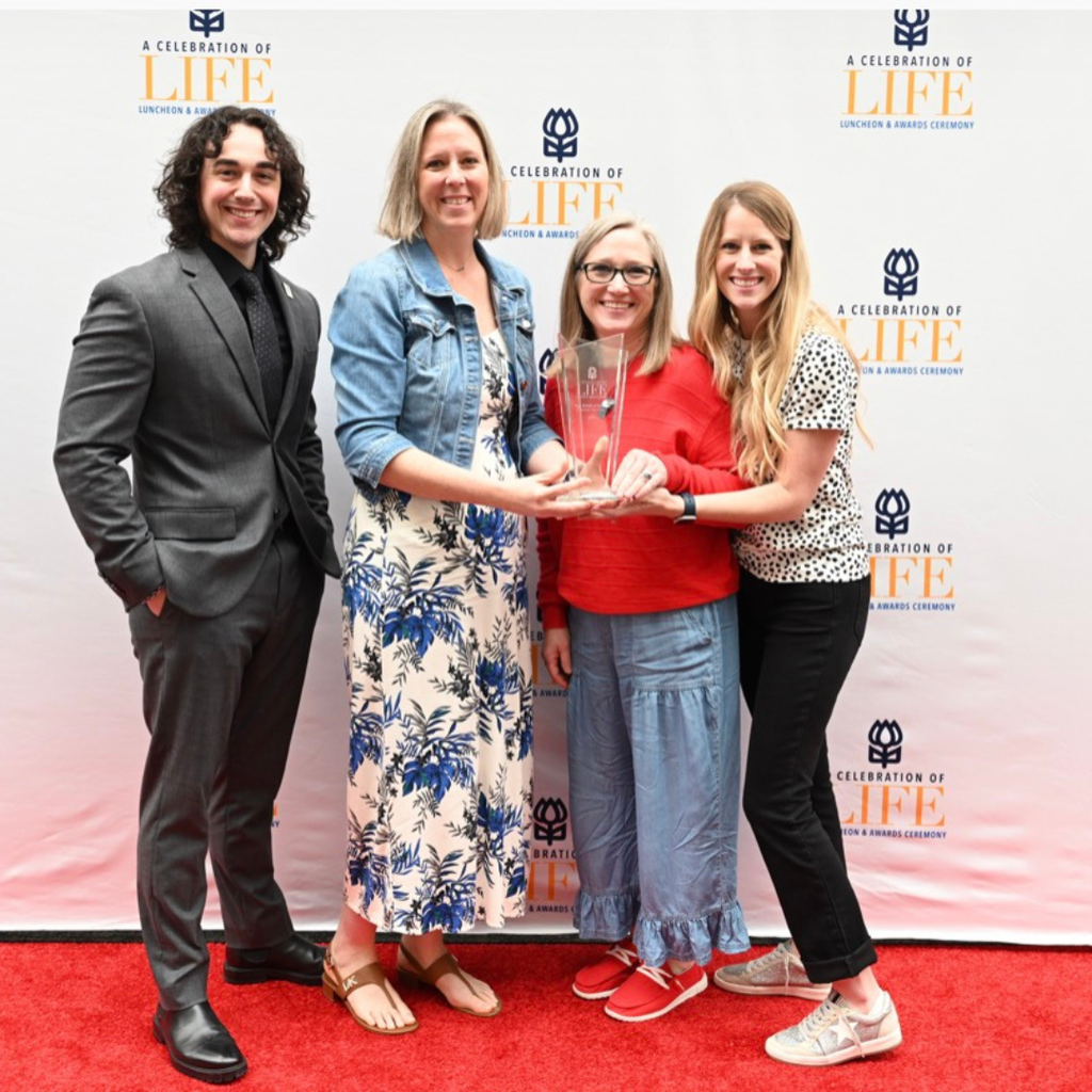 3 teachers holding a trophy posing with a man in a suit at the blood bank luncheon