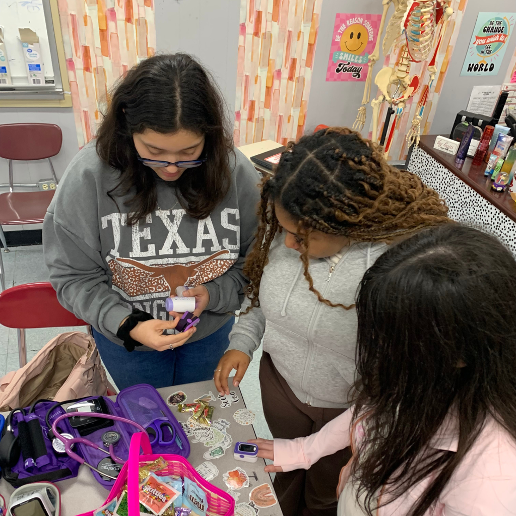 Three female students in a classroom setting focus intently on a pulse oximeter and other medical supplies on a table.
