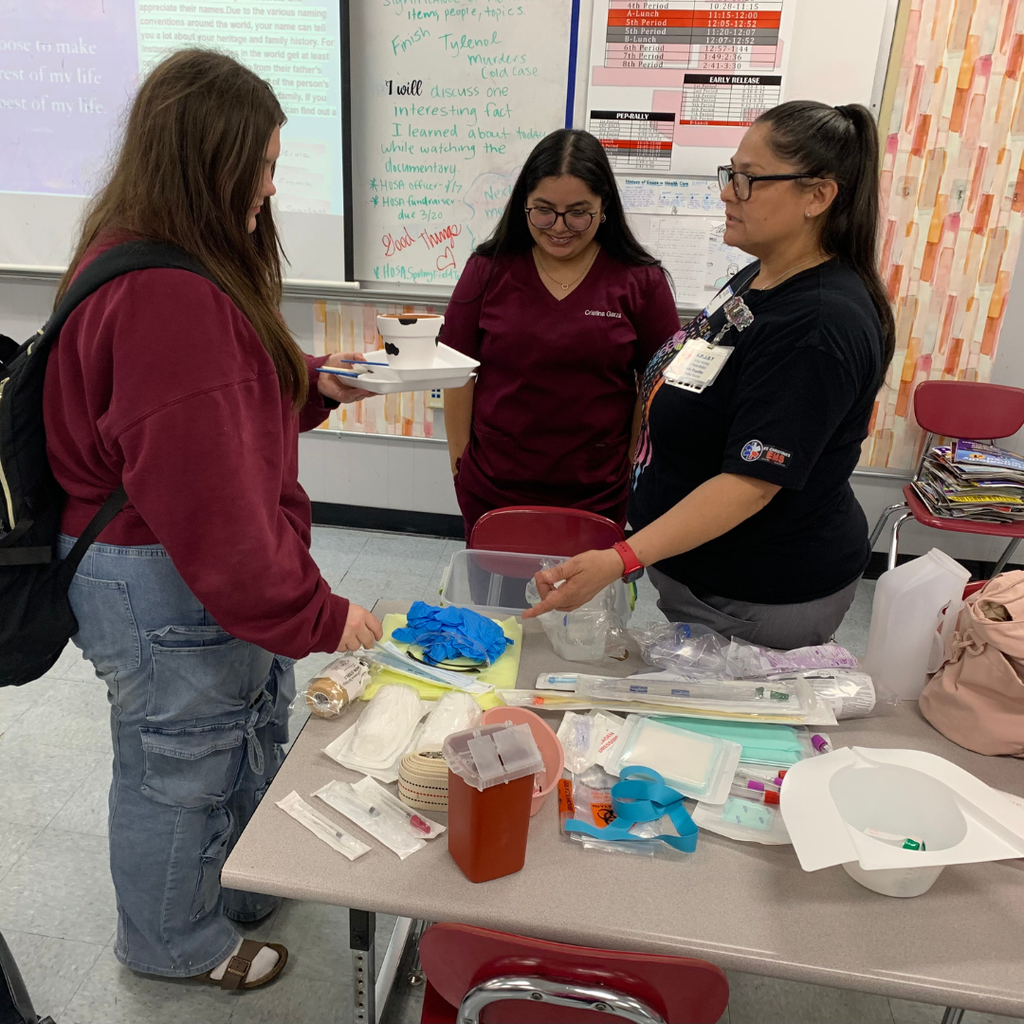A guest speaker in a "Nurse" shirt demonstrates medical supplies to two high school students at a table filled with healthcare equipment like bandages and a sharps container.