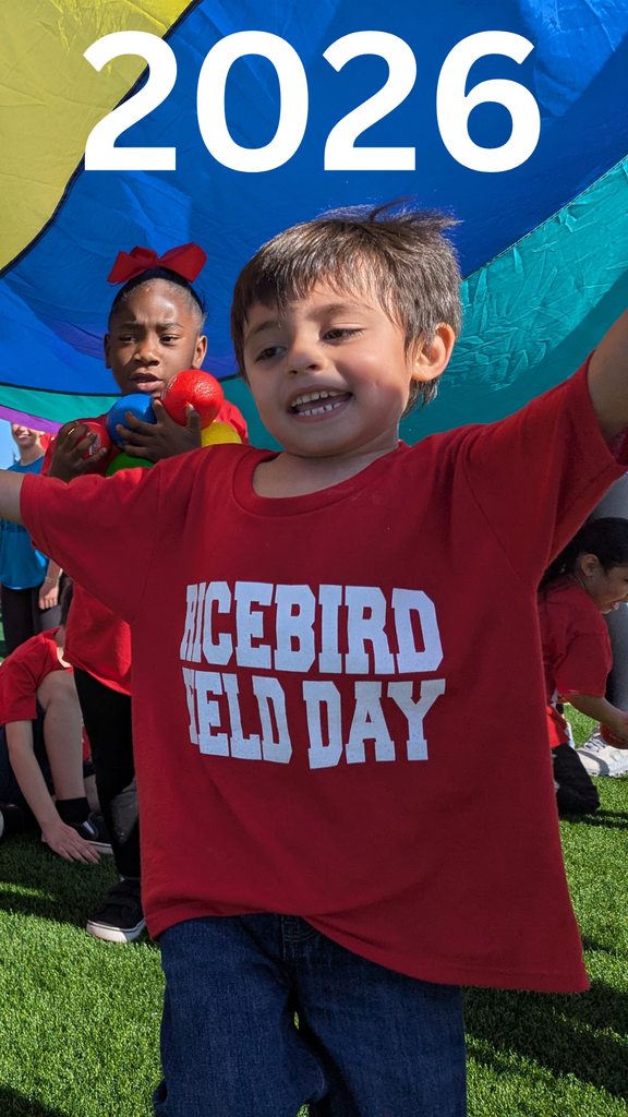 A young boy in a red "Ricebird Field Day" t-shirt smiles brightly in the foreground while other students and a teacher hold up a large, colorful rainbow parachute on a sunny day at a turf athletic field.