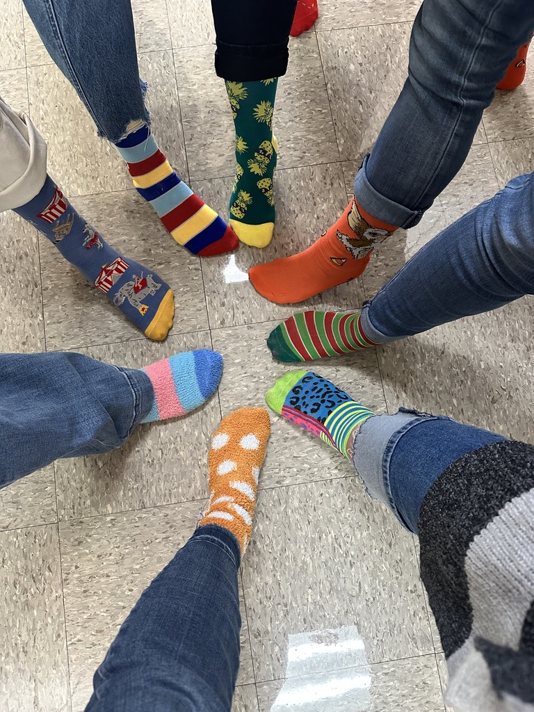 A top-down view of seven people standing in a circle, showcasing their bright, mismatched, and colorful patterned socks to celebrate World Down Syndrome Day. The socks include various designs like stripes, polka dots, and fun illustrations, worn with blue jeans against a neutral tiled floor.