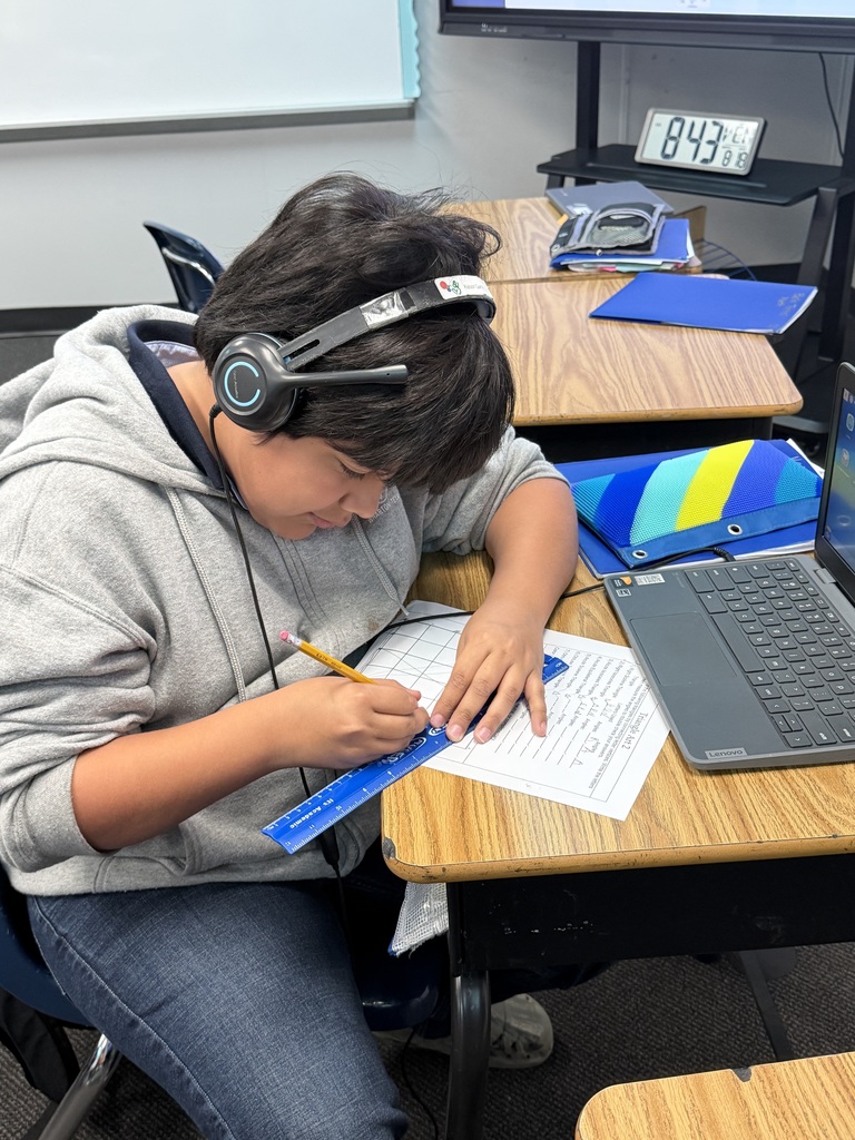 A student wearing large black headphones sits at a school desk, using a blue ruler and a pencil to carefully draw straight lines on a "Triangle Art" worksheet. A laptop is visible on the desk in front of them.