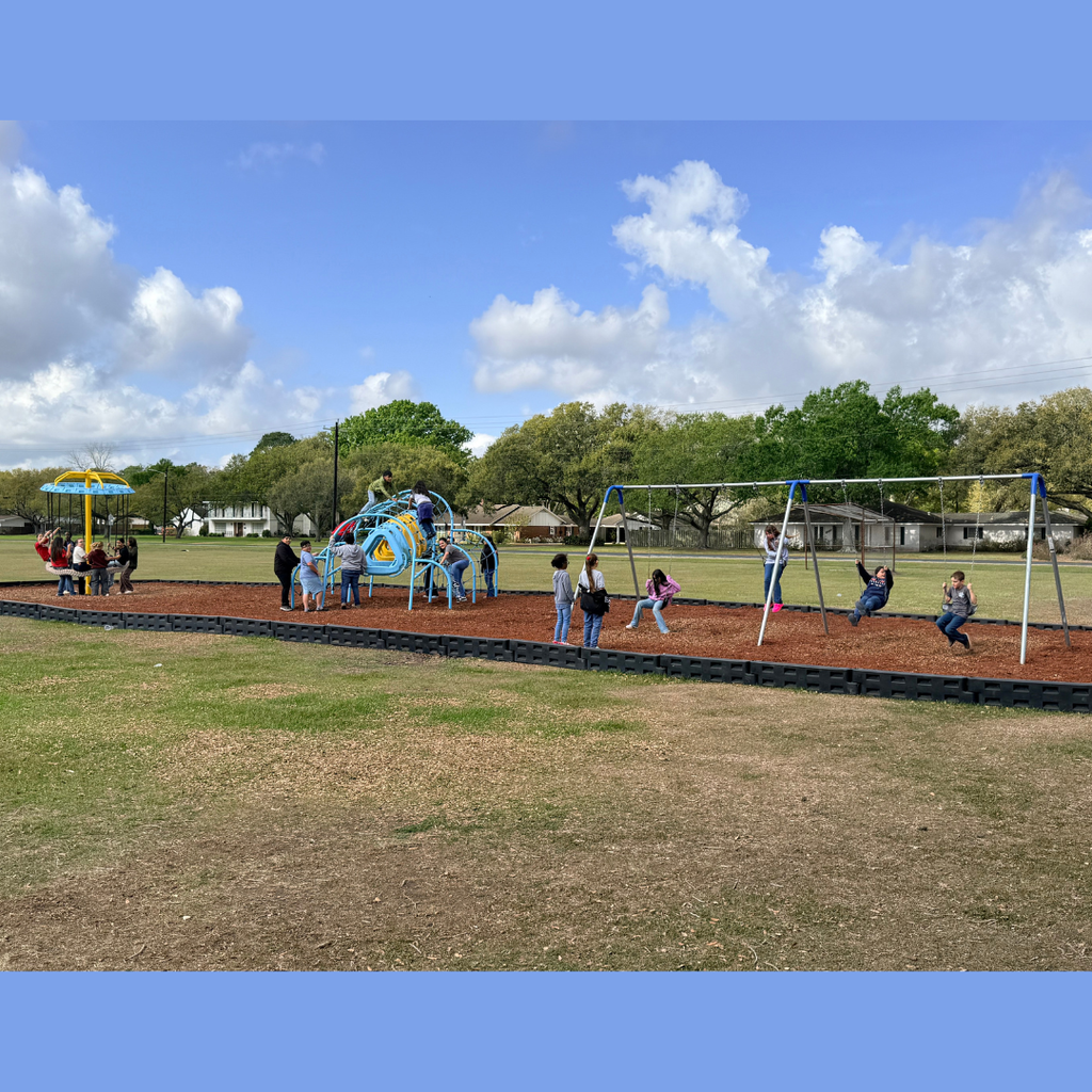 Students playing on a variety of playground equipment, including a blue climbing structure, giant tire swing, and a swing set, on a grassy field under a bright, cloudy sky