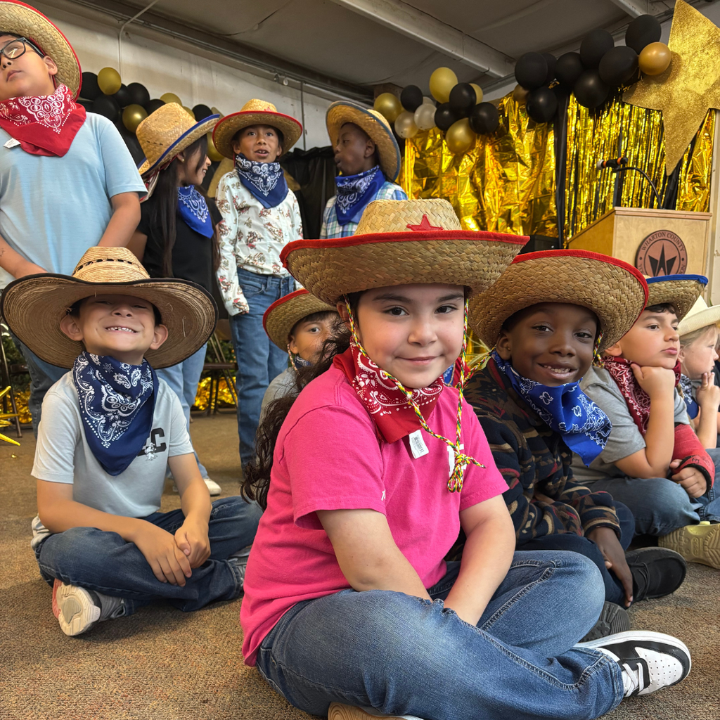 2nd-grade students from Hutchins Elementary smiling for a photo; they are wearing various straw cowboy hats with colorful bandanas, seated in front of a festive gold and black balloon display.