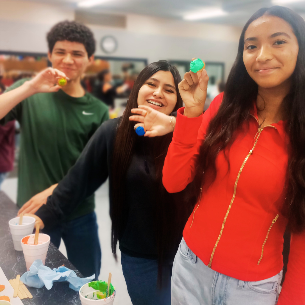A boy and two girls in a classroom setting, smiling while holding up dollops of bright green and blue frosting; they are practicing color-blending techniques for their "cookie color wheel" project.