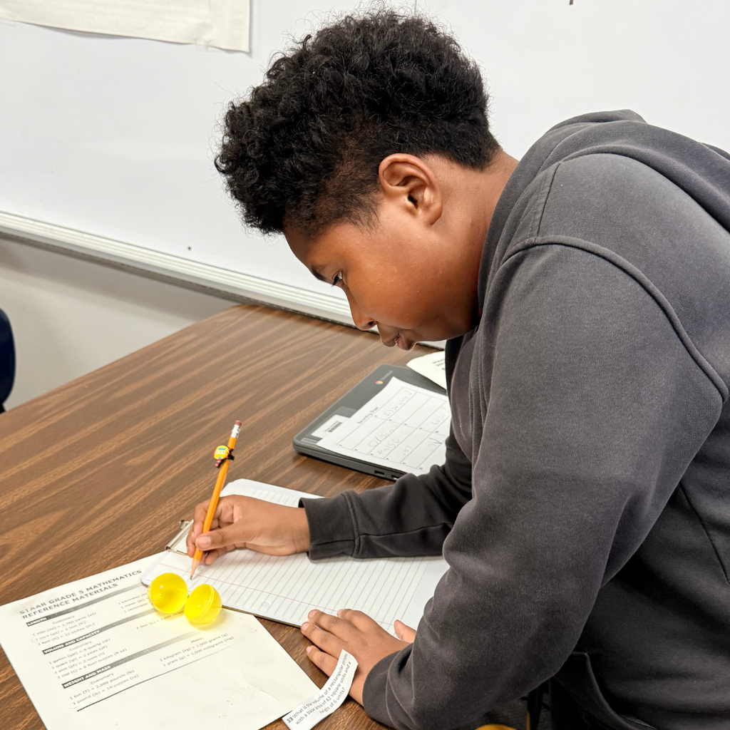 A student in a grey hoodie sitting at a desk and using a pencil to write on a math worksheet next to a yellow plastic egg.