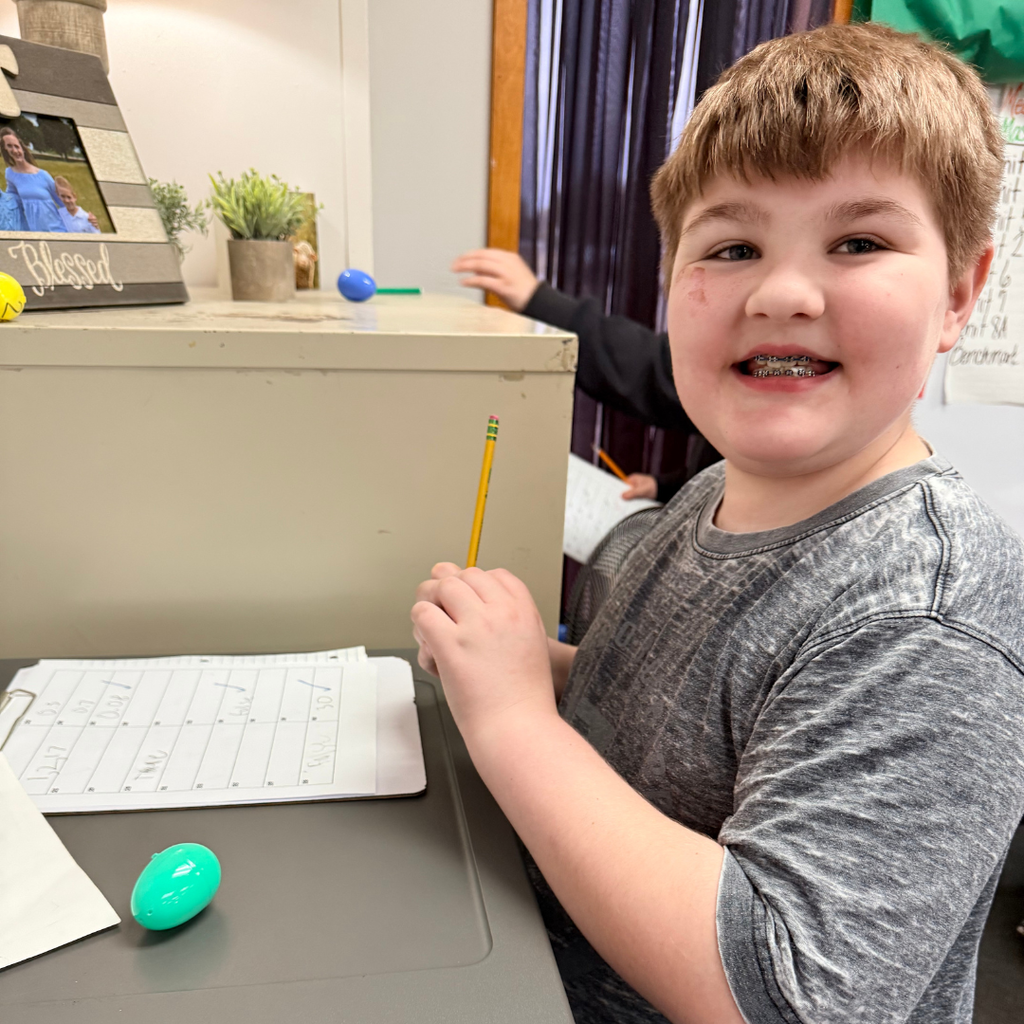 A smiling student with braces sitting at a classroom desk with a pencil and a teal plastic Easter egg.