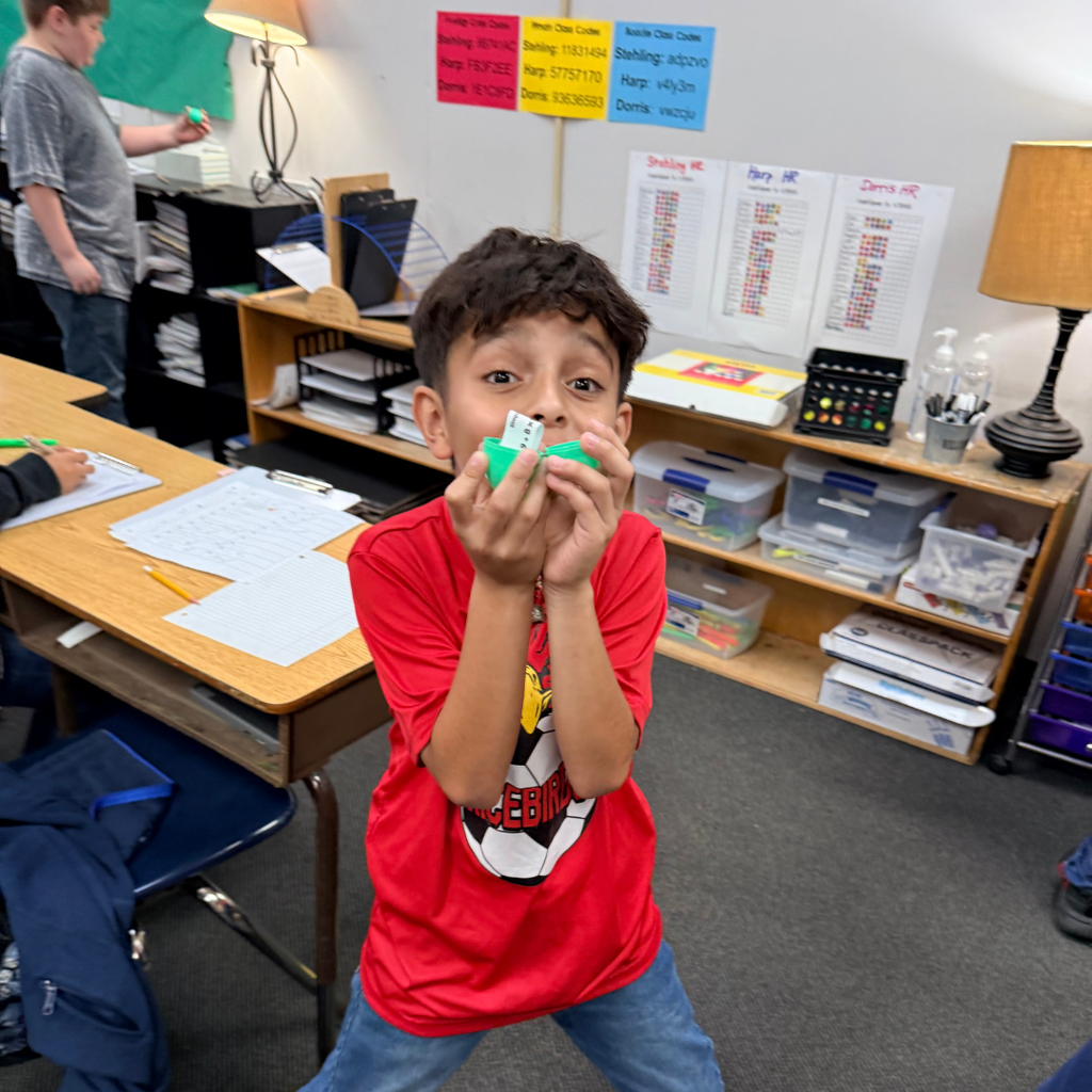 A young boy in a red shirt excitedly holds a small green Easter egg toward the camera. In the background, other students work at their desks in a brightly decorated classroom with educational posters on the walls.