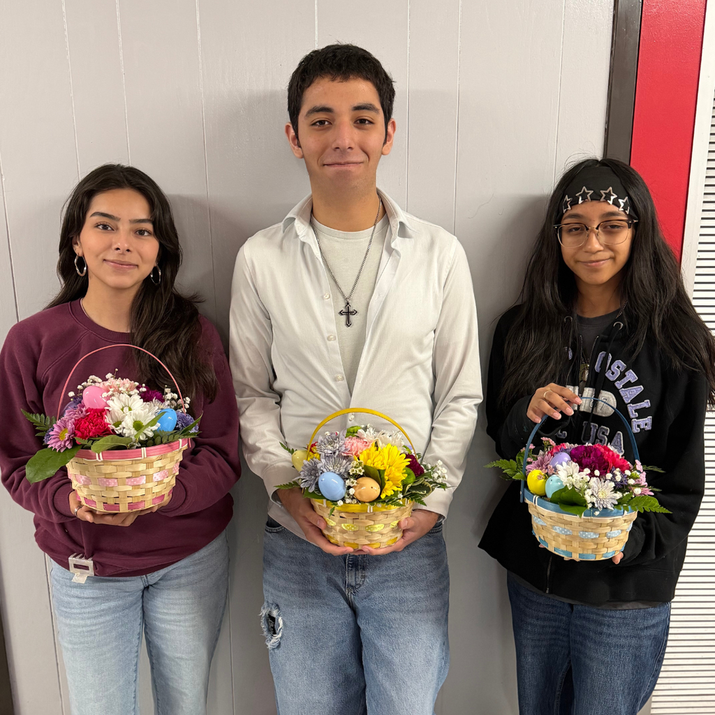 students holding an intricate floral arrangement featuring a variety of spring flowers, greenery, and pastel ribbons in a wicker basket, displayed on a classroom table.