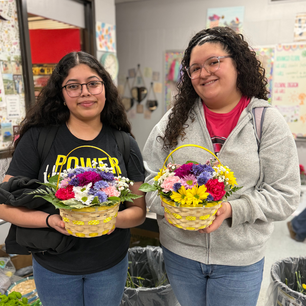 students holding an intricate floral arrangement featuring a variety of spring flowers, greenery, and pastel ribbons in a wicker basket, displayed on a classroom table.