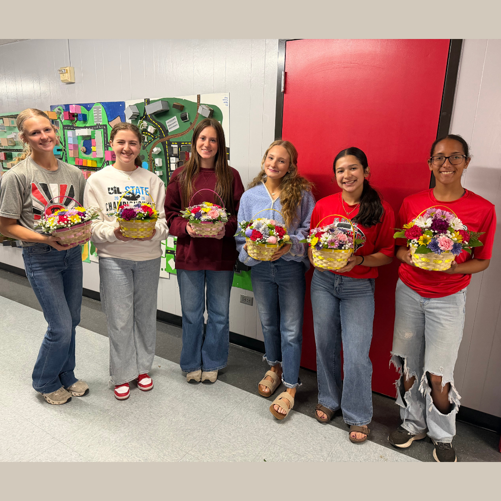 students holding an intricate floral arrangement featuring a variety of spring flowers, greenery, and pastel ribbons in a wicker basket, displayed on a classroom table.