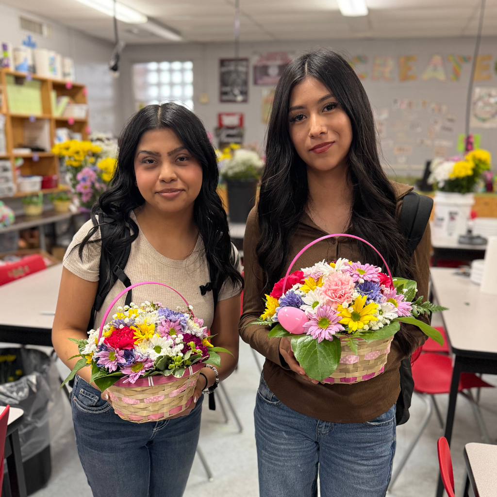 students holding an intricate floral arrangement featuring a variety of spring flowers, greenery, and pastel ribbons in a wicker basket, displayed on a classroom table.