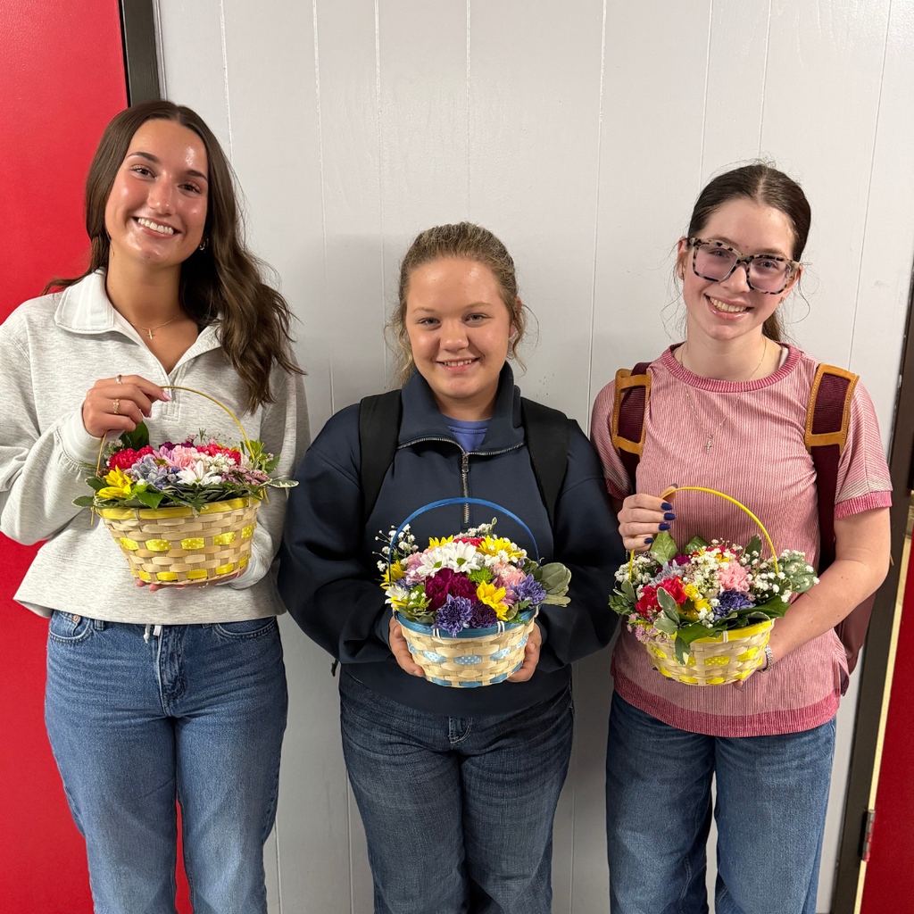 students holding an intricate floral arrangement featuring a variety of spring flowers, greenery, and pastel ribbons in a wicker basket, displayed on a classroom table.