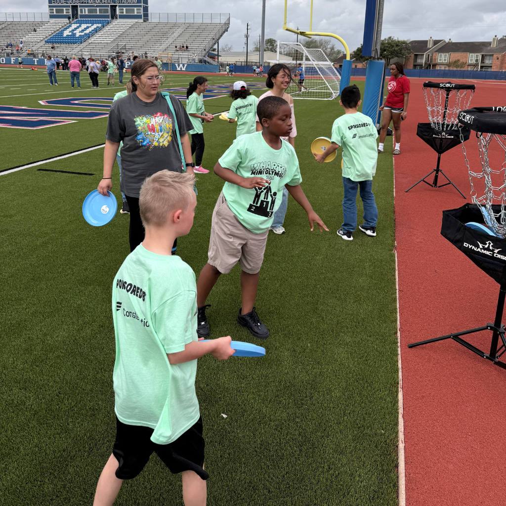 elementary students in light green t-shirts participating in the Wharton Mini Olympics. The children are outdoors on a track and field, smiling and playing with blue flying discs and sports equipment alongside their teachers.