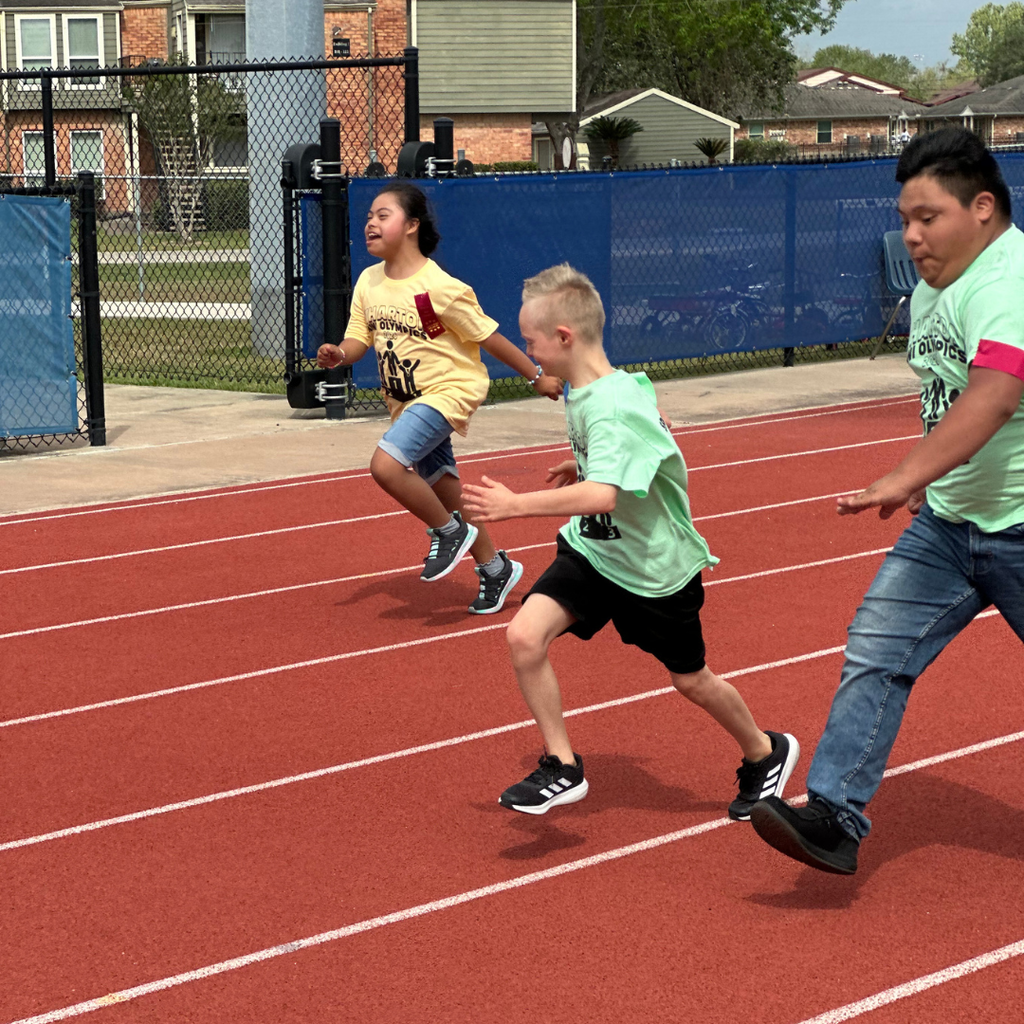 Elementary student in light green t-shirt participating in the Wharton Mini Olympics outdoors on a track, smiling and running alongside other kids.