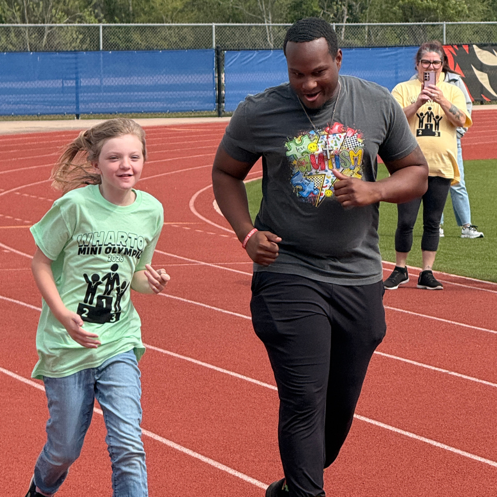 elementary student in light green t-shirt participating in the Wharton Mini Olympics outdoors on a track, smiling and running alongside a teacher.